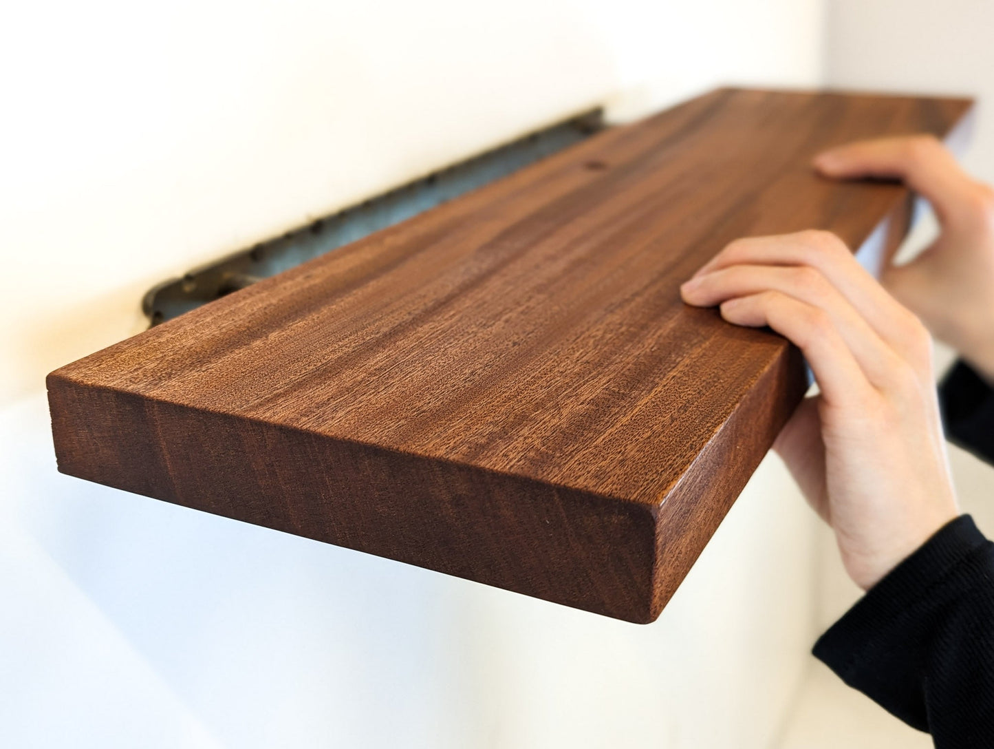 Two hands push a mahogany shelf with a grommet hole onto a metal bracket that is attached to the wall.