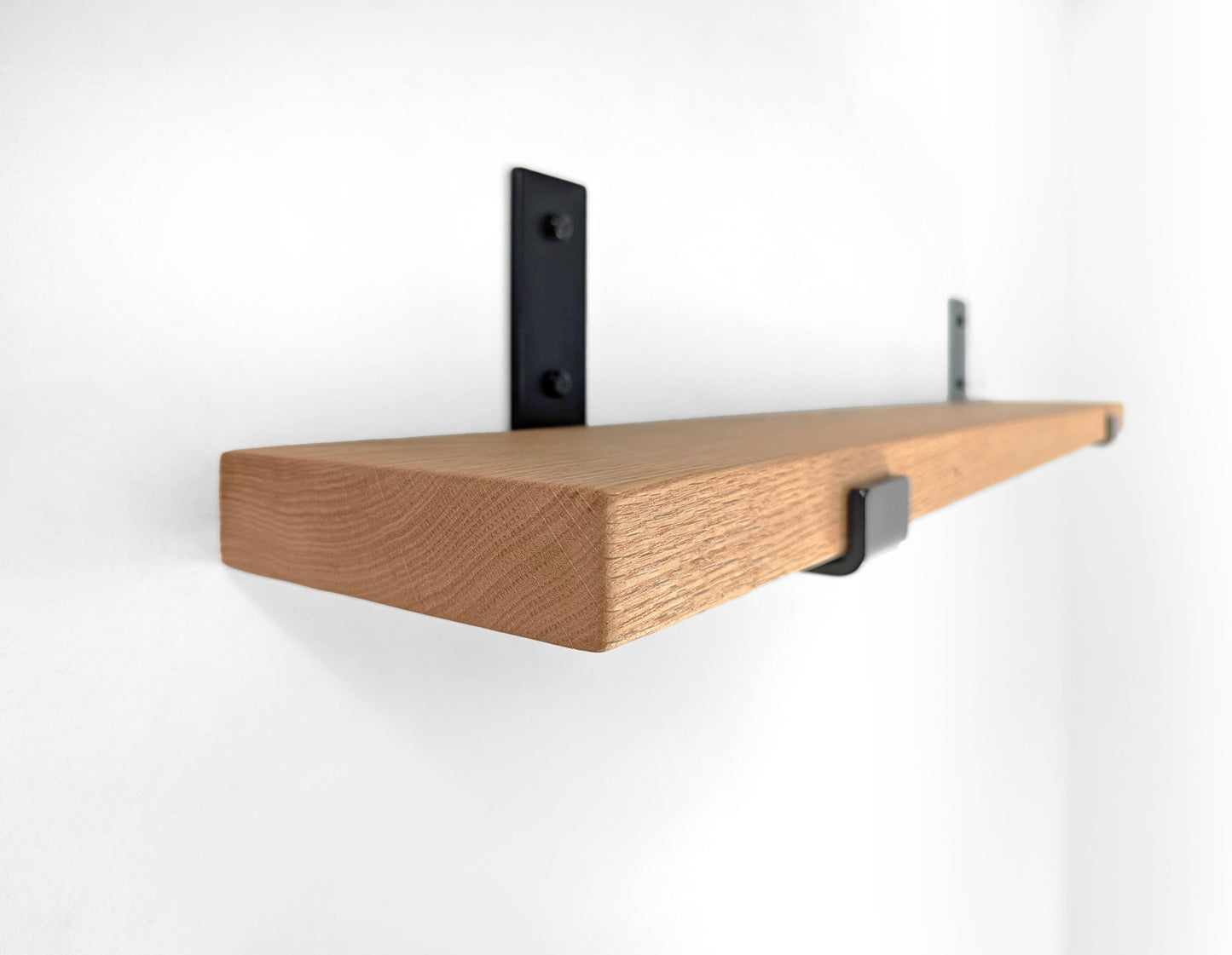 Side angle view of a long oak shelf with two J brackets attached to the wall with bolts.