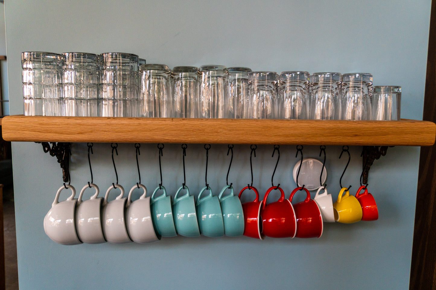 A high-quality and customizable coffee mug shelf is mounted on a blue wall. Various sized glasses are stacked on top of the shelf and below hang five white mugs, four blue mugs, three red mugs, and a yellow and orange espresso cups. In the back, out of focus is a bar tap with seven wooden taps in descending sizes.