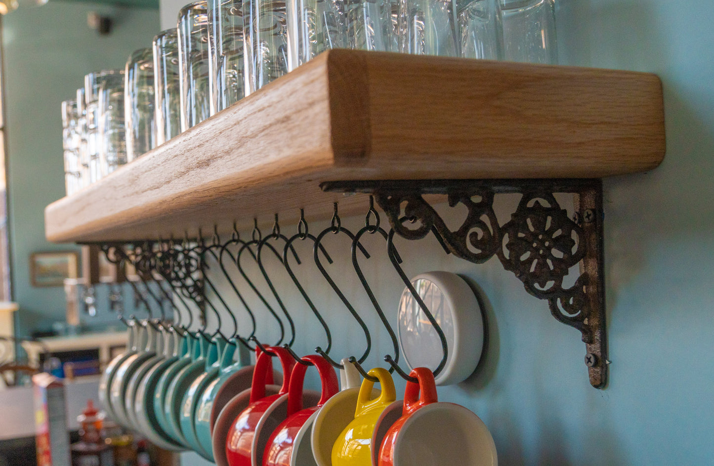 A high-quality and customizable coffee mug shelf is mounted on a blue wall. Various sized glasses are stacked on top of the shelf and below hang five white mugs, four blue mugs, three red mugs, and yellow and orange espresso cups.
