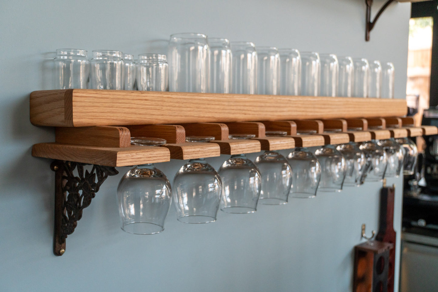 A side view of our Oak or Mahogany Beer Glass Shelf. The oak shelf holds 13 drink glasses securely as a variety of tulip glasses hang upside down below. A cast-iron bracket features a floral pattern. The shelf is mounted on a blue wall.