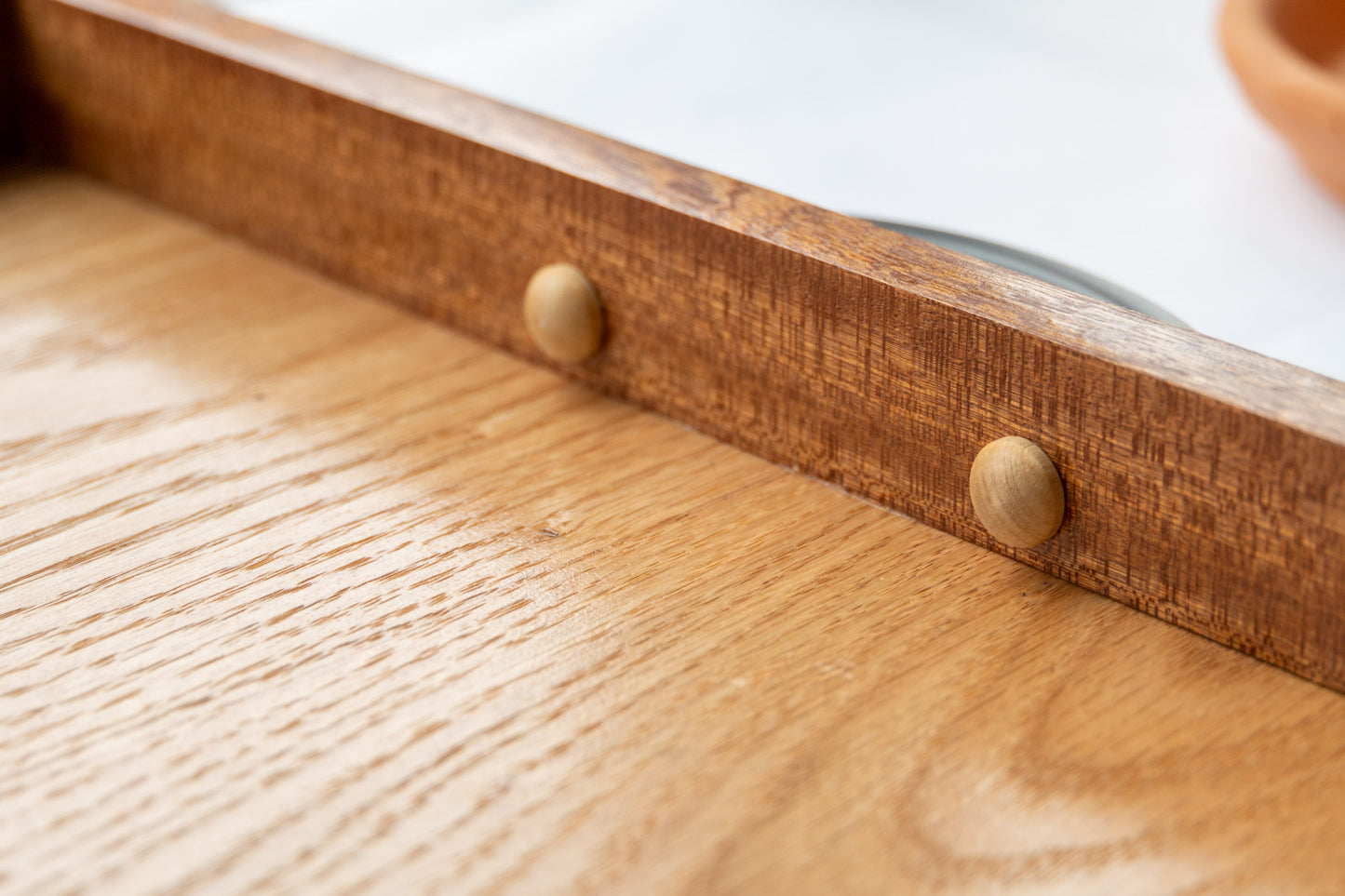 A close-up of the mahogany lip of our Luxury Serving Tray with Full Grain Leather. The dark mahogany wood elegantly meets the light oak wood that makes up the base of the tray. Two oak buttons protrude slightly on the inside of the serving tray to offer safe handling with the dark brass handles.