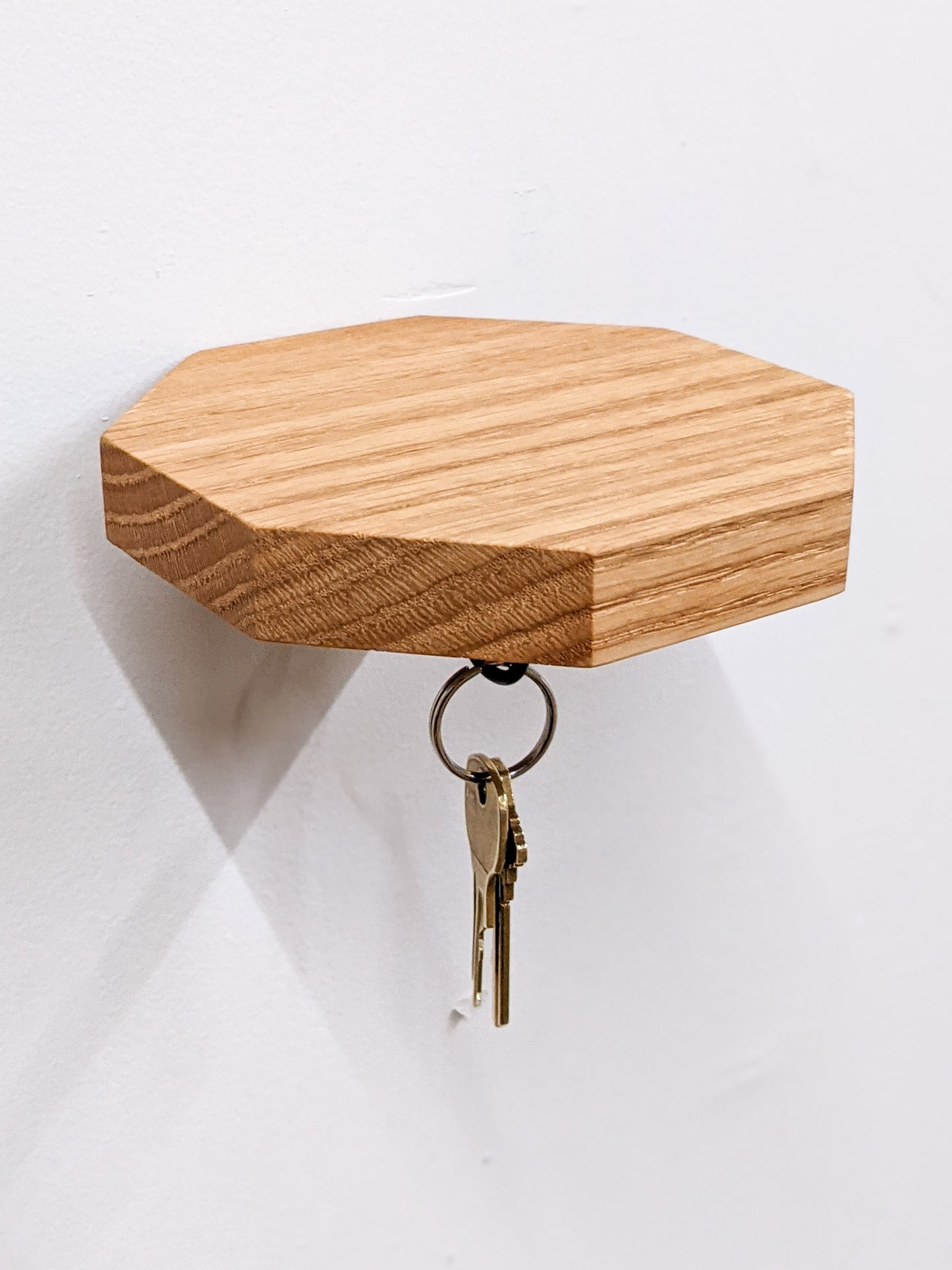 A floating oak octagon key shelf has been successfully installed on a white wall. Two bronze keys dangle from a black key hook on the bottom. The top is empty allowing for an excellent view of the natural fine grain of the oak wood to shine. 