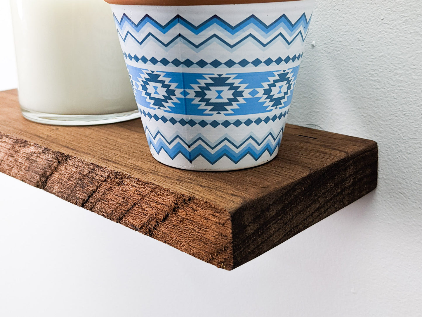 A medium mahogany live edge floating wall shelf is wall mounted and holds two planters that are cut off at the top of the picture. The far left planter is pure white and the planter to the right is white and blue.