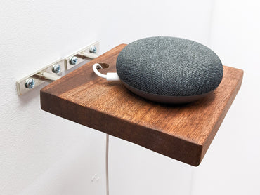 A small round speaker sits on top of a small mahogany square floating shelf that is mid-installation. A wire is plugged into the speaker and easily slips through a drilled hole in the shelf for easy and stylish cord management.