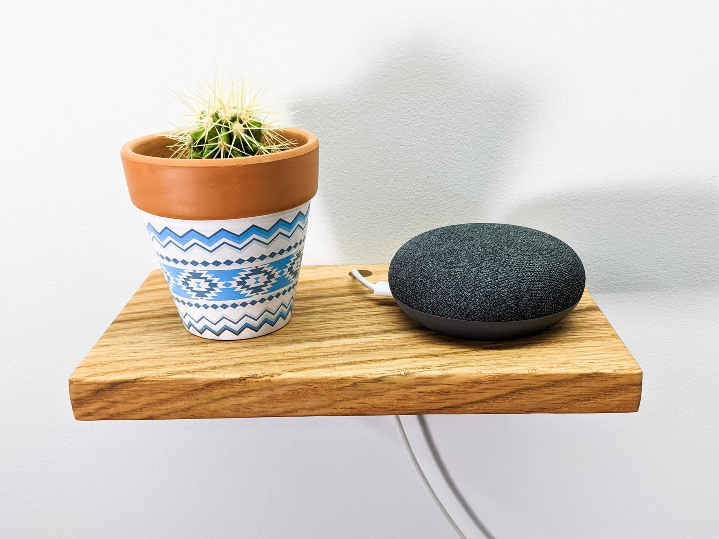 A rectangular floating wall shelf holds a round corded speaker and unique white and blue planter with a round cactus inside of it. The white cord of the speaker seamlessly slips through a drilled hole in the back and blends into the white wall.