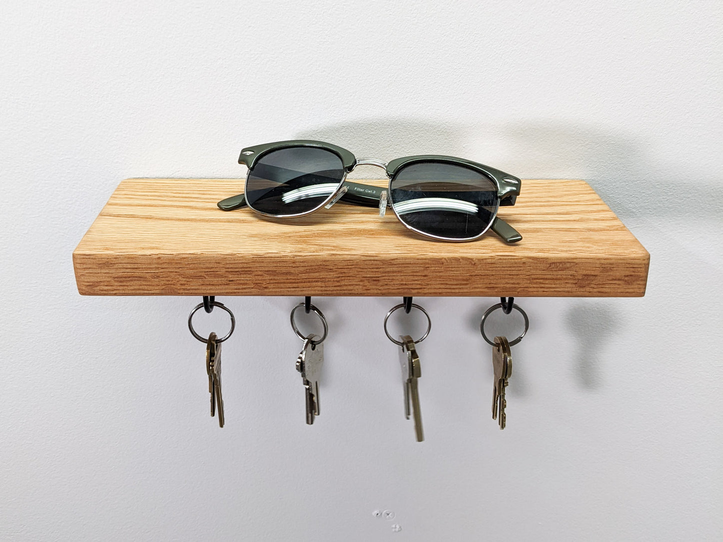 A head-on view of NookWoodworking's oak key hook shelf. The soft fine grain of the oak wood displays natural whirls on the wall-mounted base. A pair of black sunglasses rest on top. Below, four hanging black key hooks hold eight keys. Four keys are bronze and four are silver. 