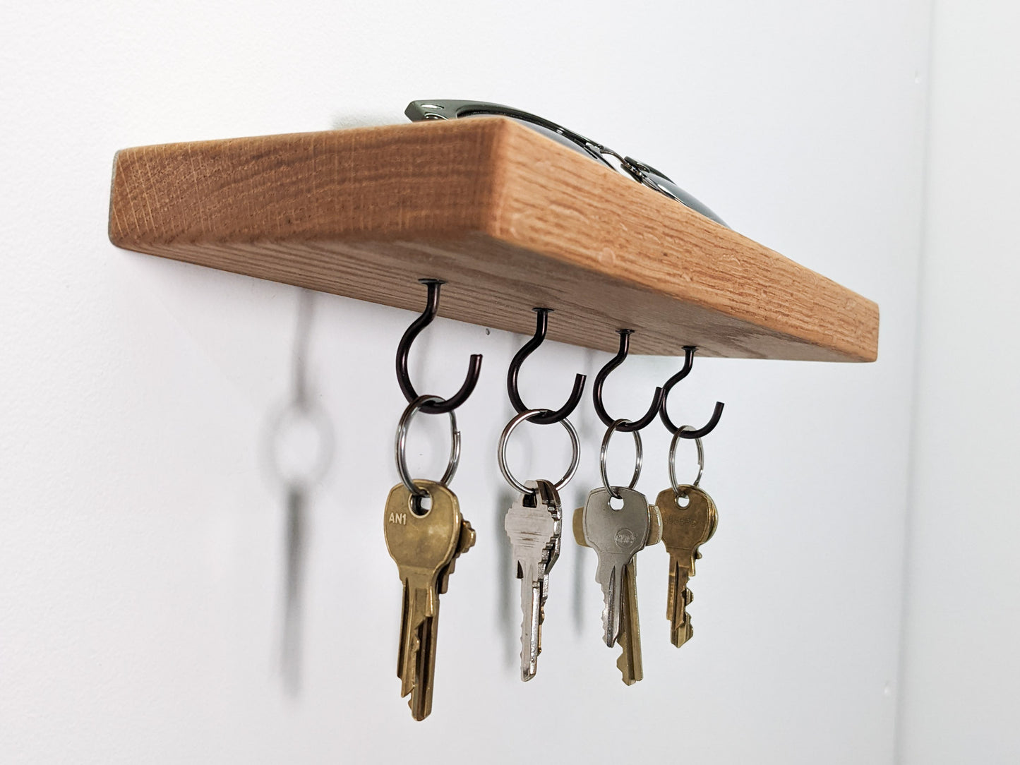Four black key hooks hang securely from a wall-mounted floating oak shelf . Four keys are bronze, four are silver. The keys cast a slight shadow onto the white wall behind them. On top of the shelf sits a pair of sunglasses.