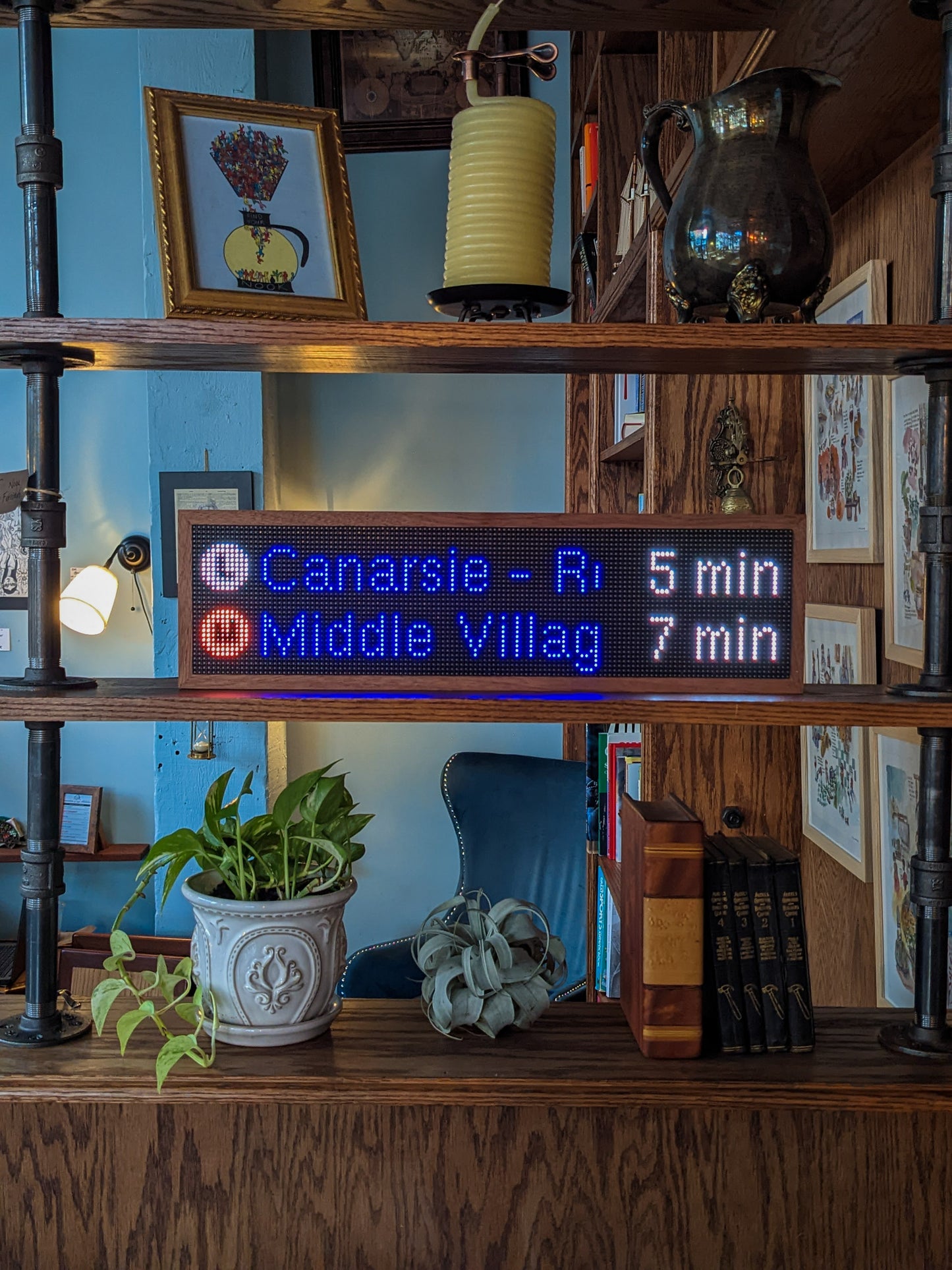 Three shelves of a mahogany bookcase are on display. The top shelf holds an illustration of a coffee carafe, wick candle, and antique pitcher, the middle shelf holds the subway clock with mahogany frame. The subway clock reads, "L Canarsie -R 5 min" and below that, "M Middle Villag 7 Min", on the bottom shelf a green plant sits in an intricate white planter next to a silver air plant, a large brown book, and four black books. The wall behind it is light blue. There is a blue accent chair in the back.