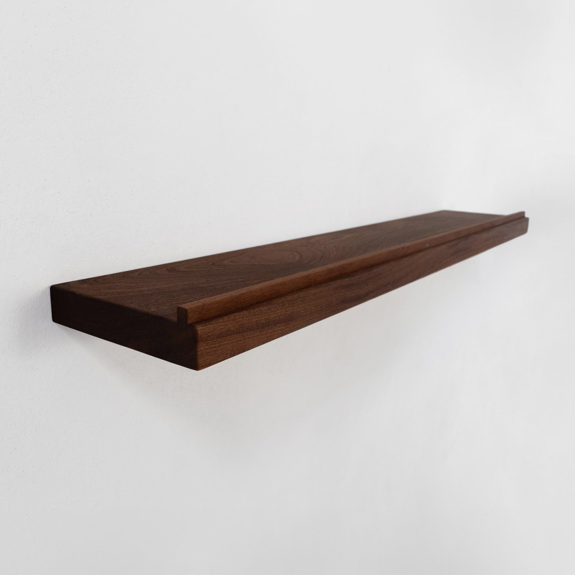 A long, minimalist floating wooden shelf made of dark brown wood with a lip near the front edge, mounted on a plain white wall.