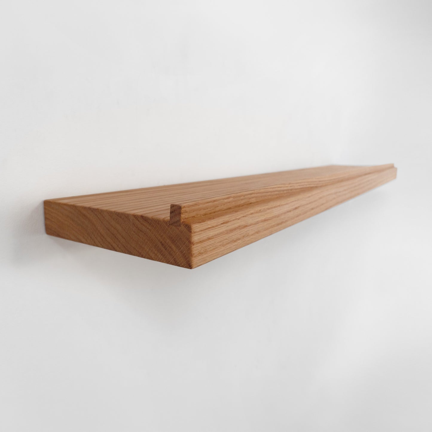 A long, minimalist floating wooden shelf made of medium brown wood and with a lip ledge at the front, shown mounted on a plain white wall.