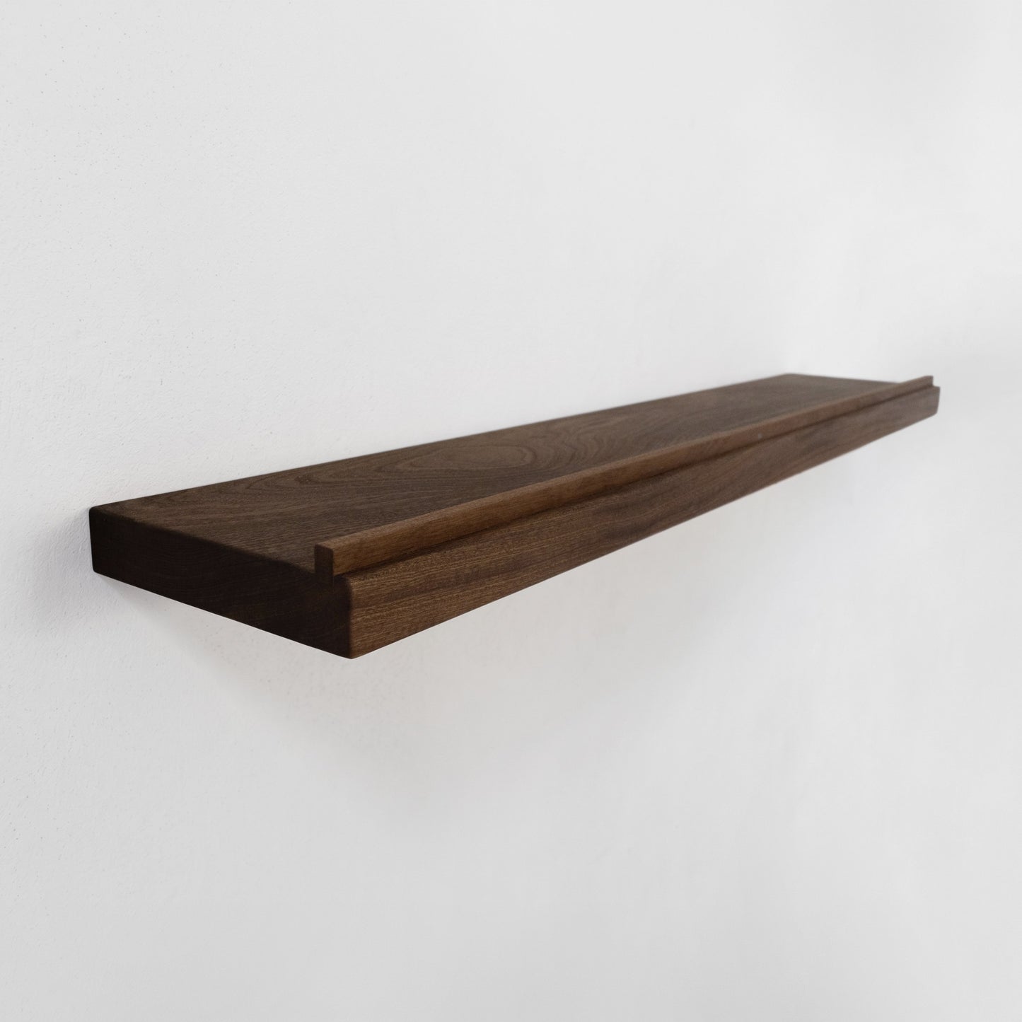 A long, dark brown, walnut, wooden floating shelf with a modern cantilevered design and a lip at the front, mounted on a plain white wall.