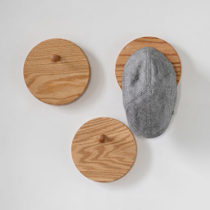 Three round wooden oak peg hooks with small knobs are mounted on a light gray wall. One hook on the right holds a gray textured cap, showing how the product is used.