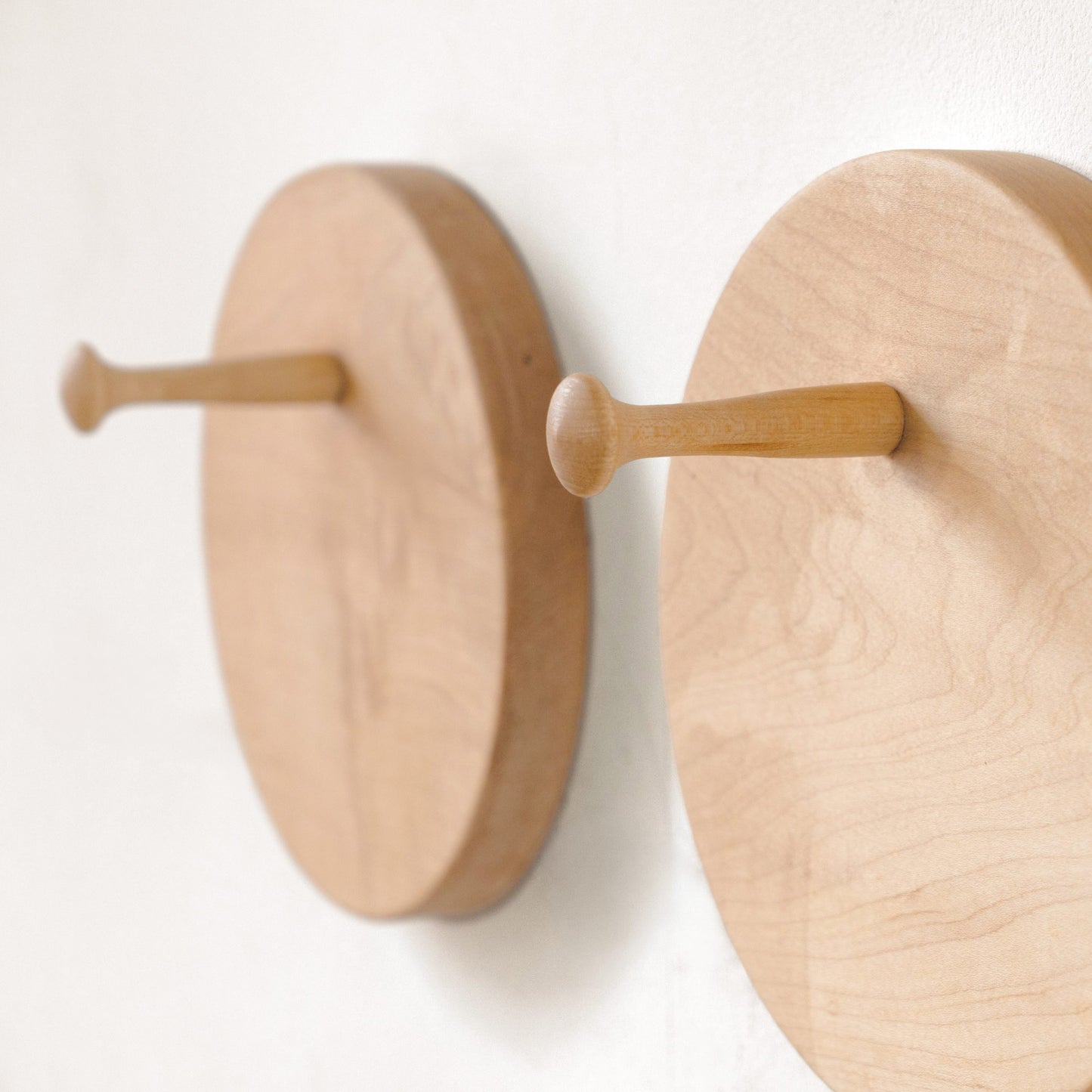 Two round light wood hat racks mounted on a white wall, shown in a close-up with one hook in front of the other.