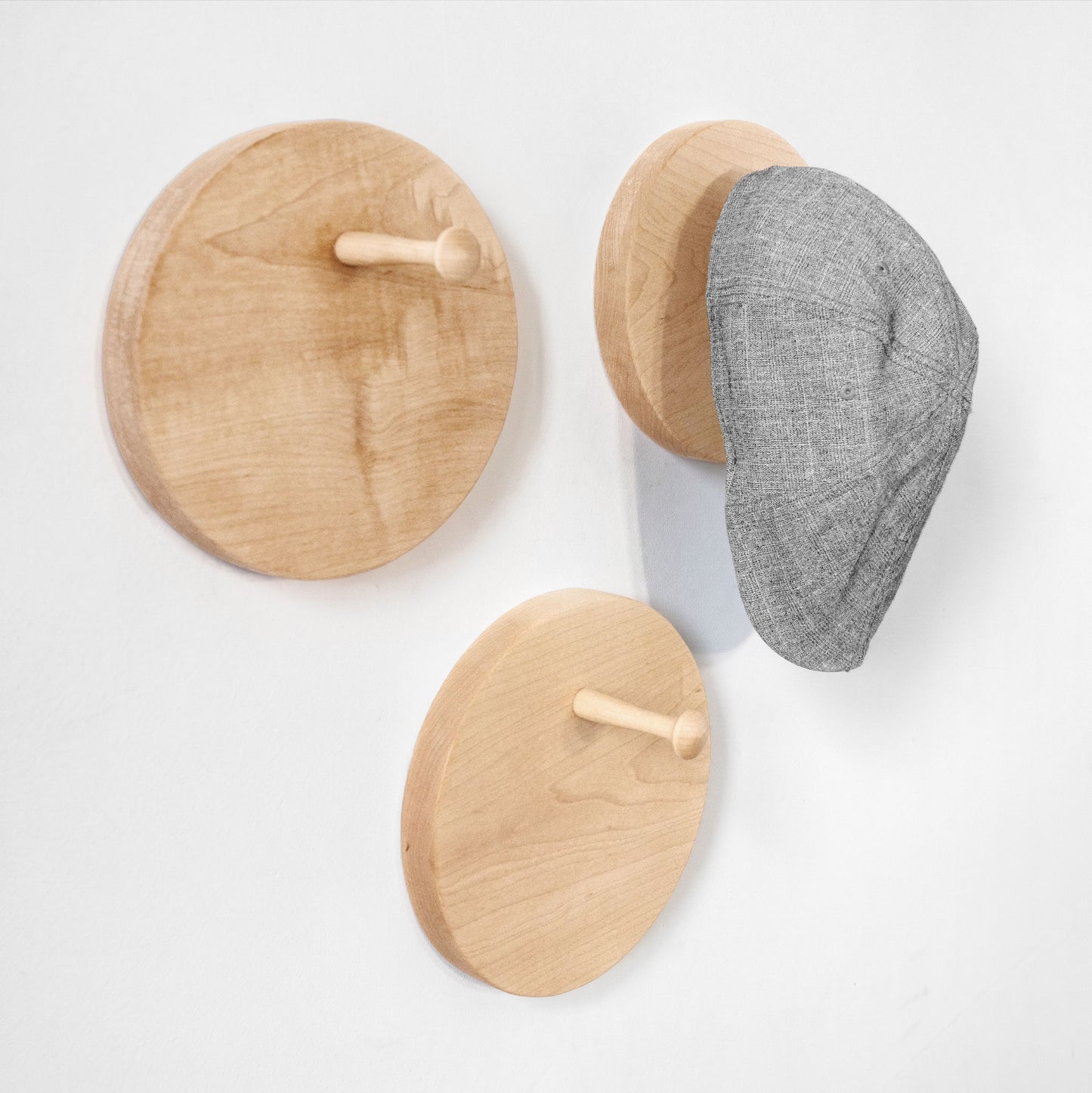 Three round wooden peg wall hat racks in light natural wood, shown on a white wall with a gray textured cap hanging on one hook.
