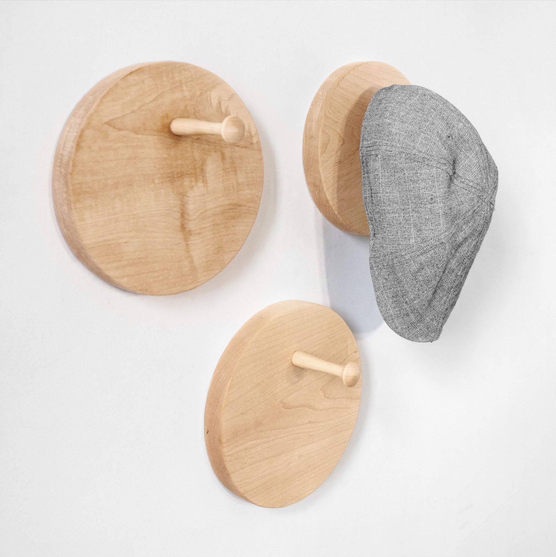 Three round wooden peg wall hat racks in light natural wood, shown on a white wall with a gray textured cap hanging on one hook.