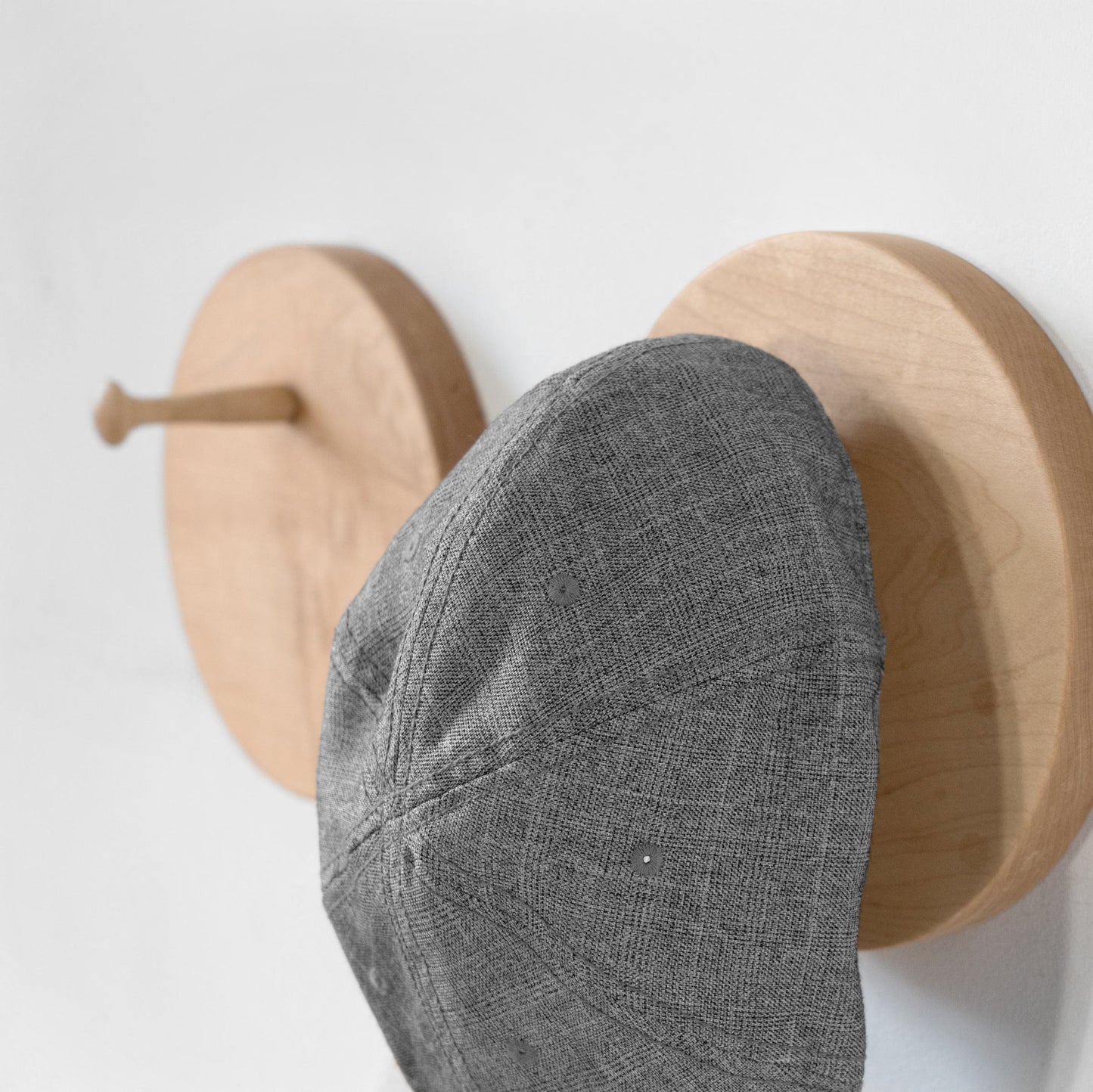 A gray textured flat cap hanging from a light wood wall-mounted hat rack hook on a white wall.