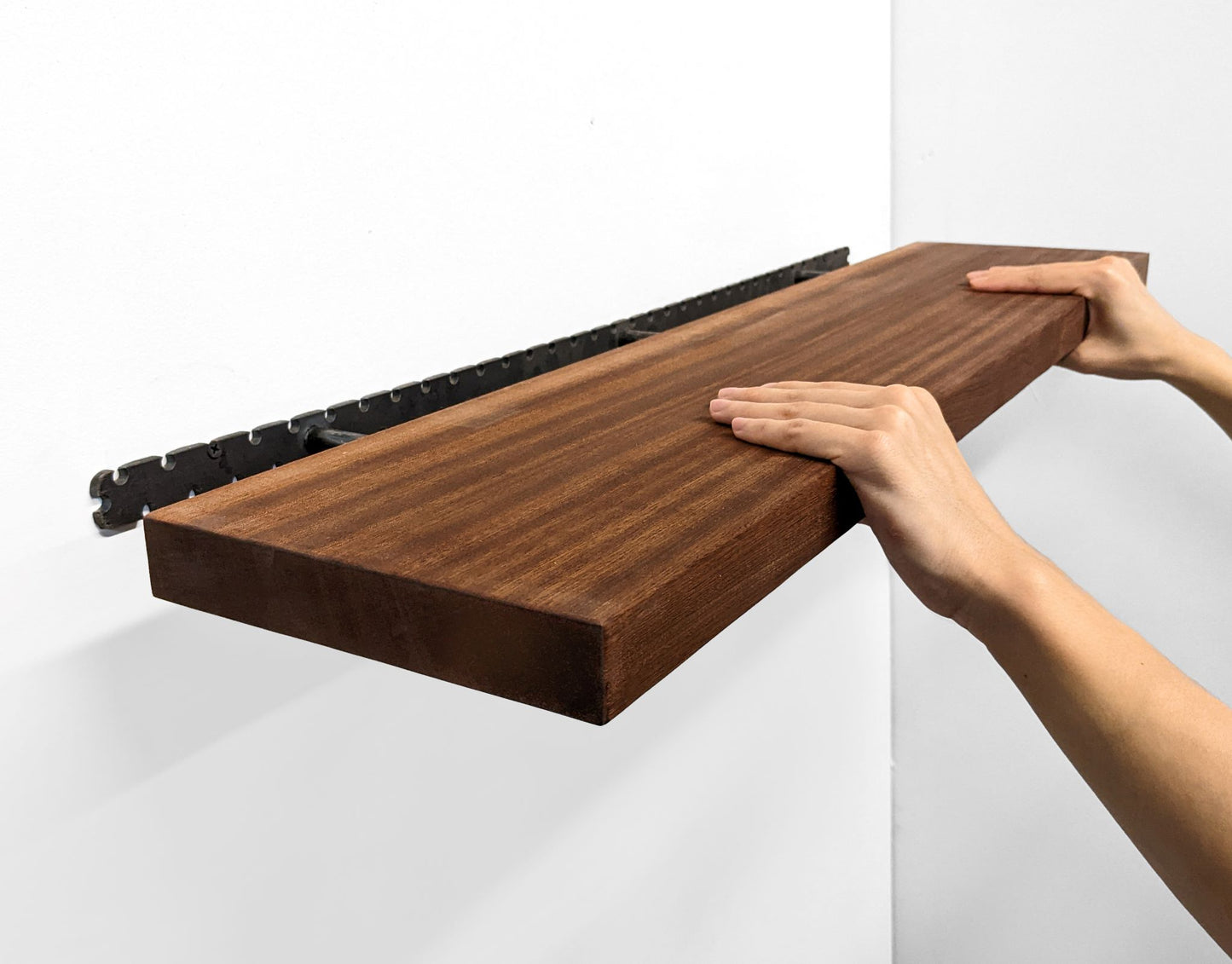 Two hands grip the front of a long wooden mahogany shelf and easily install it on the wall by sliding it onto black brackets.