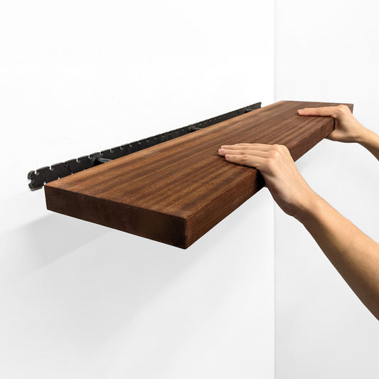 Two hands grip the front of a long wooden mahogany shelf and easily install it on the wall by sliding it onto black brackets.
