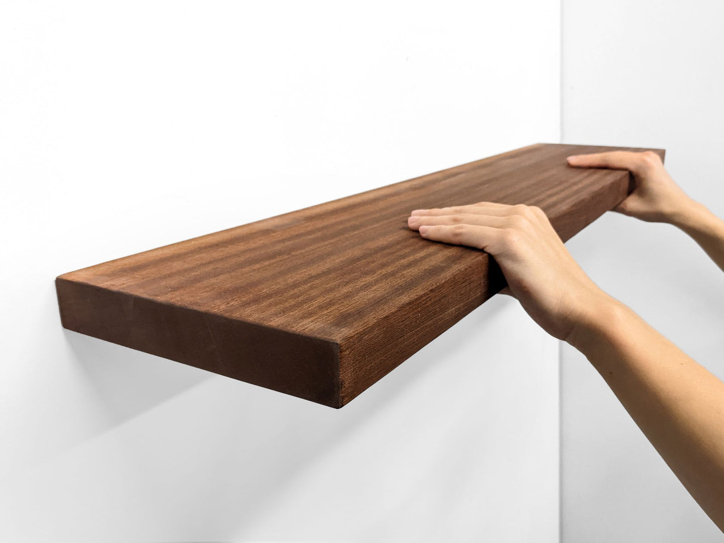A long floating shelf floats on a wall. The brown mahogany wood displays the natural grain of the heavy duty shelf is highlighted. The shelf casts a shadow on the wall.