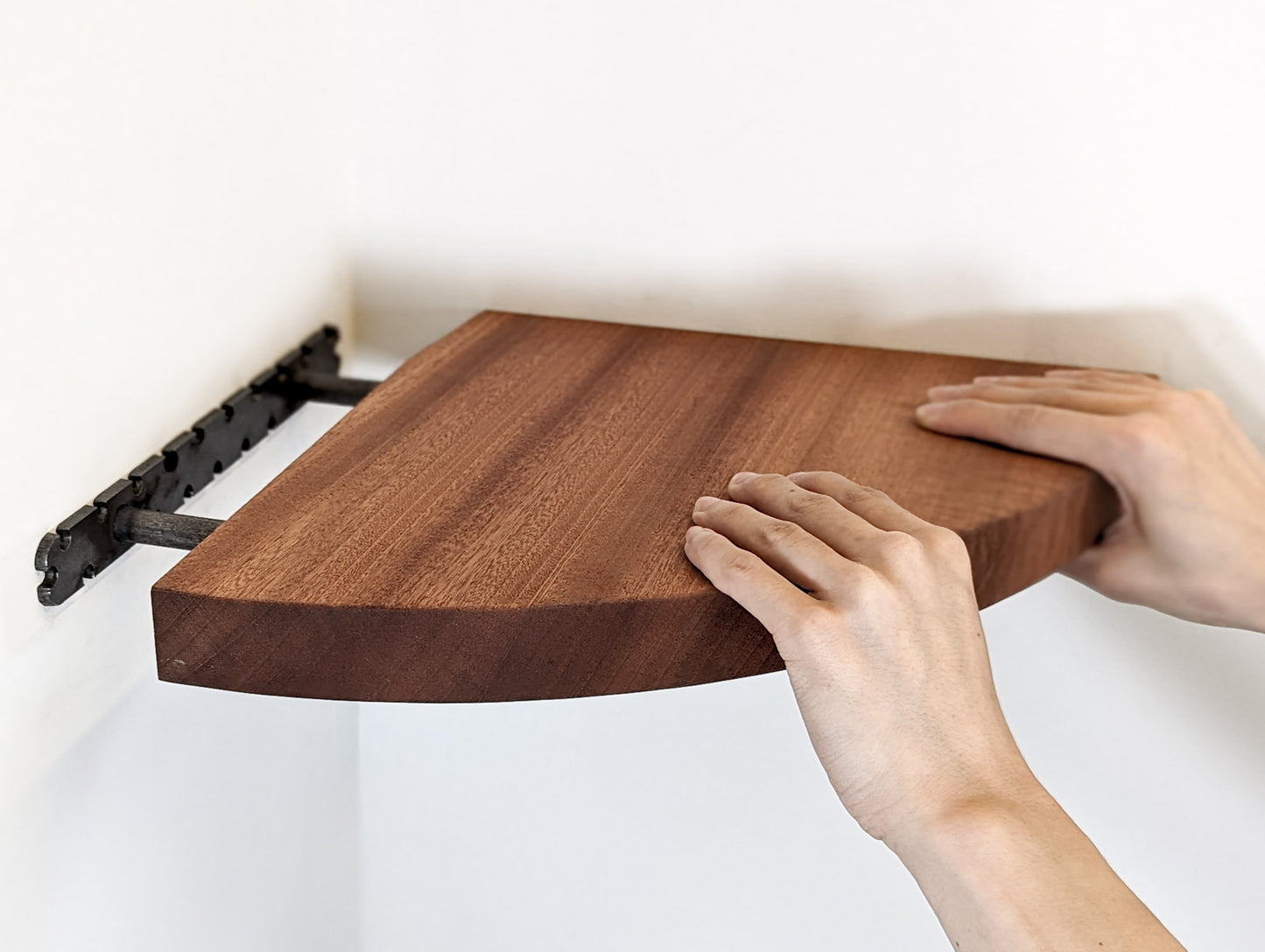 Two hands push a mahogany corner shelf onto the brackets which are wall-mounted.