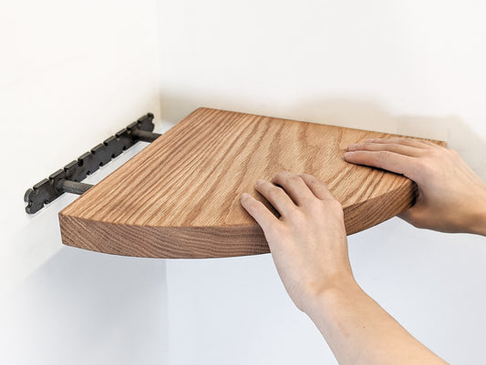 Two hands push a solid wood oak floating corner shelf onto the bracket.