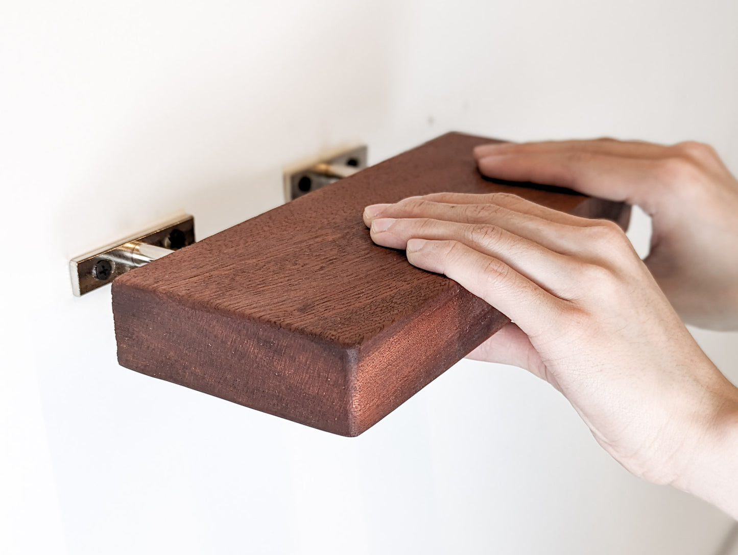 Two hands slide a mahogany shelf on to it's two metal brackets that are attached to the wall.