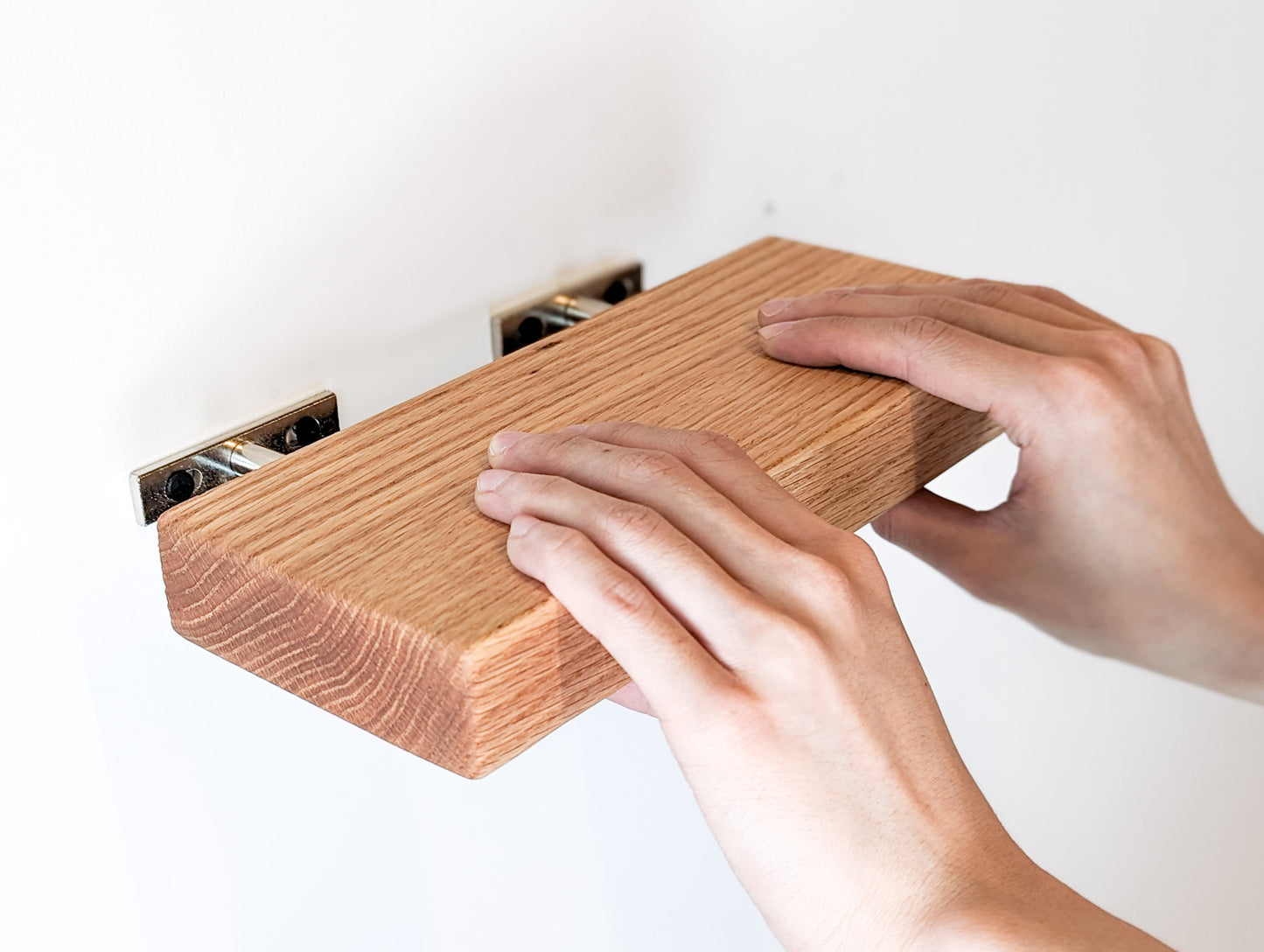 Two hands push an oak shelf onto the metal brackets that are attached to the wall.