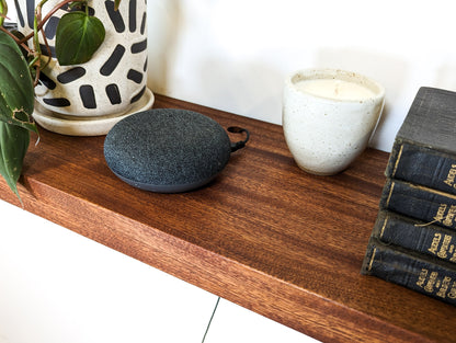 A speaker cord is shown through the grommet hole on a mahogany shelf. A candle, four books, and a plant are also displayed on the shelf.