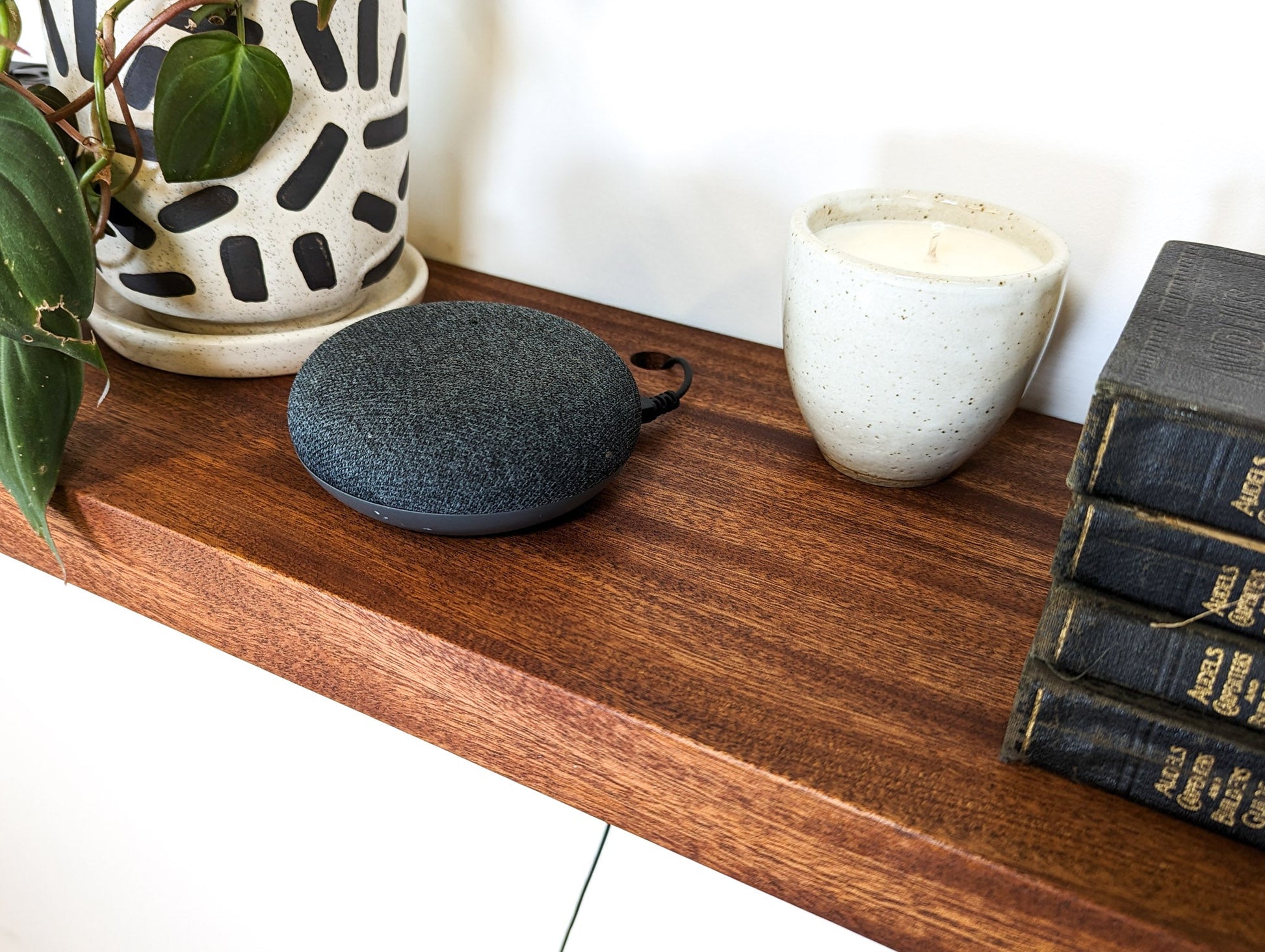 A speaker cord is shown through the grommet hole on a mahogany shelf. A candle, four books, and a plant are also displayed on the shelf.