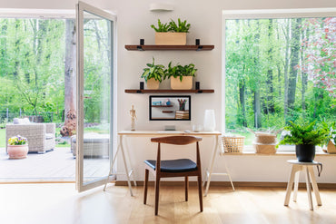 Two thick mahogany shelves with steel black powder coated J brackets are on a wall displaying plants above a desk between two windows.