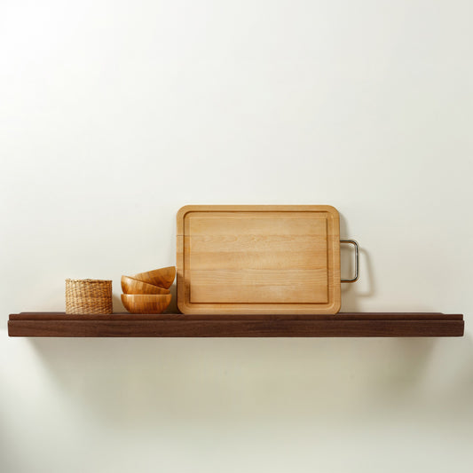 A rectangular light wood cutting board with a metal handle resting on a dark wooden floating shelf with a lip on the front edge, alongside a small woven basket and stacked wooden bowls, against a plain off-white wall.