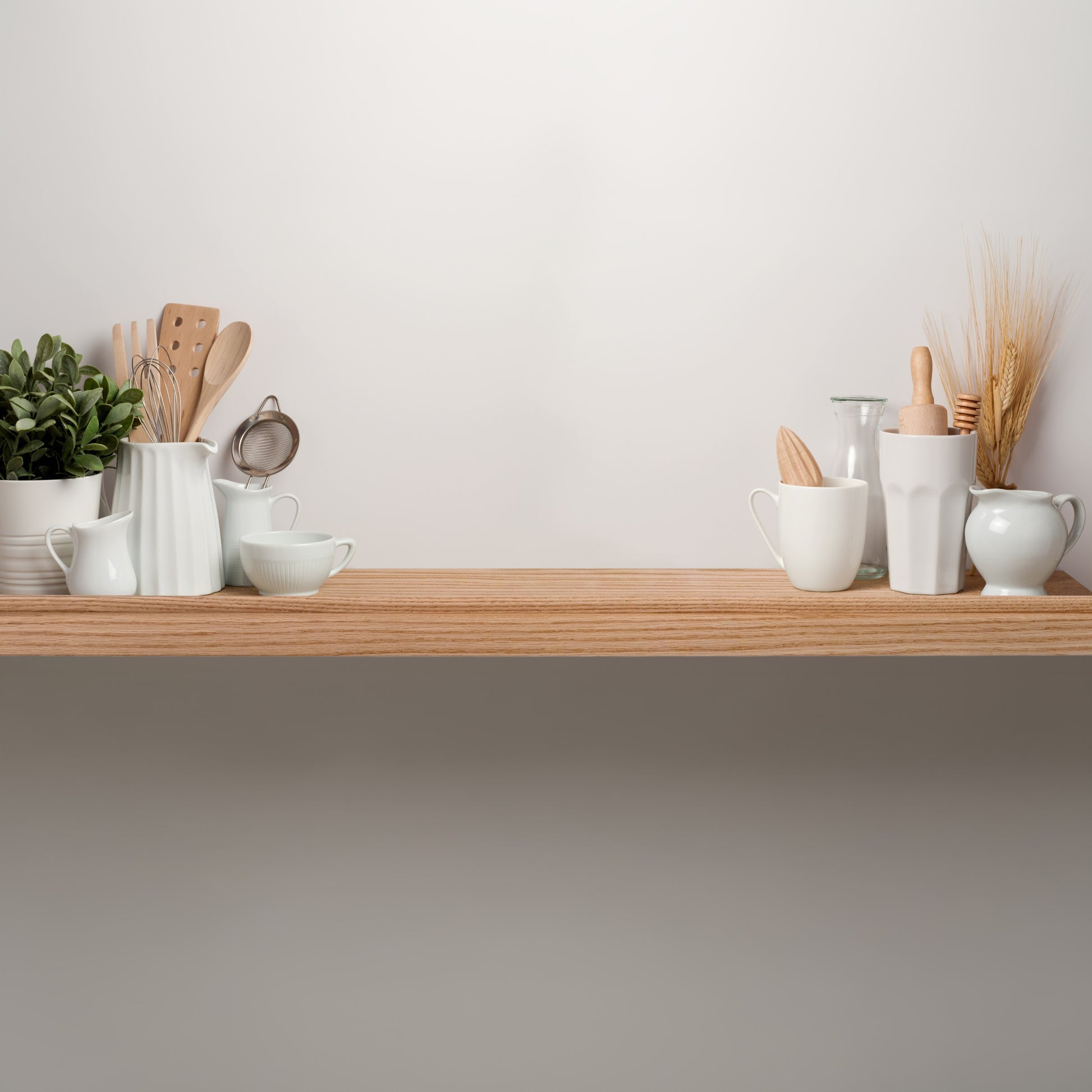 A light wood oak floating shelf with a lip near the front mounted on a white wall, styled with white ceramic kitchenware, wooden utensils, a small potted green plant, and dried wheat stalks.