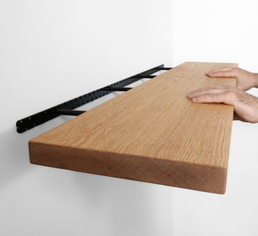 Two hands grip the top and bottom of the front piece of a long wooden oak shelf and easily install it onto the wall by sliding the shelf onto black brackets.