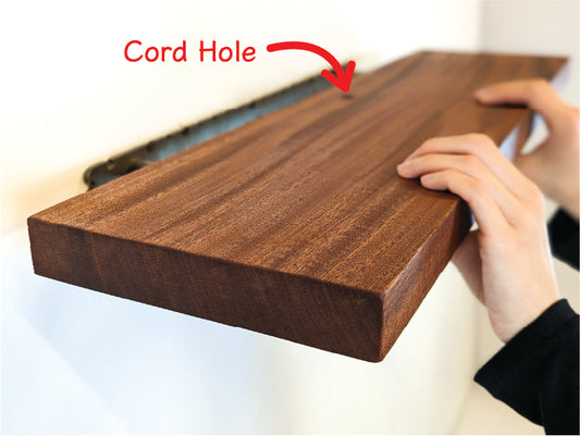 Two hands push a thick mahogany shelf with a grommet hole onto a metal bracket. Text saying "Cord Hole" points to the grommet hole in the middle of the shelf.