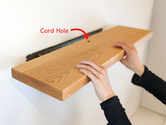 Two hands push a thick oak shelf with a grommet hole onto a metal bracket. Text is shown overlaying the image that says "Cord Hole" with an arrow to the grommet hole on the shelf.