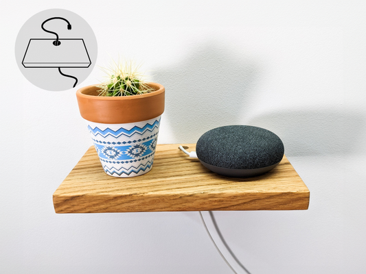 A rectangular floating wall shelf holds a round corded speaker and unique white and blue planter with a round cactus inside of it. The white cord of the speaker seamlessly slips through a drilled hole in the back and blends into the white wall. 