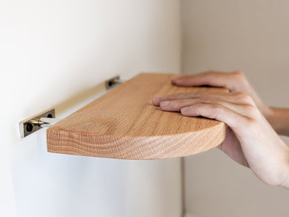 Two hands push a thin oak floating shelf with curved corners onto metal brackets