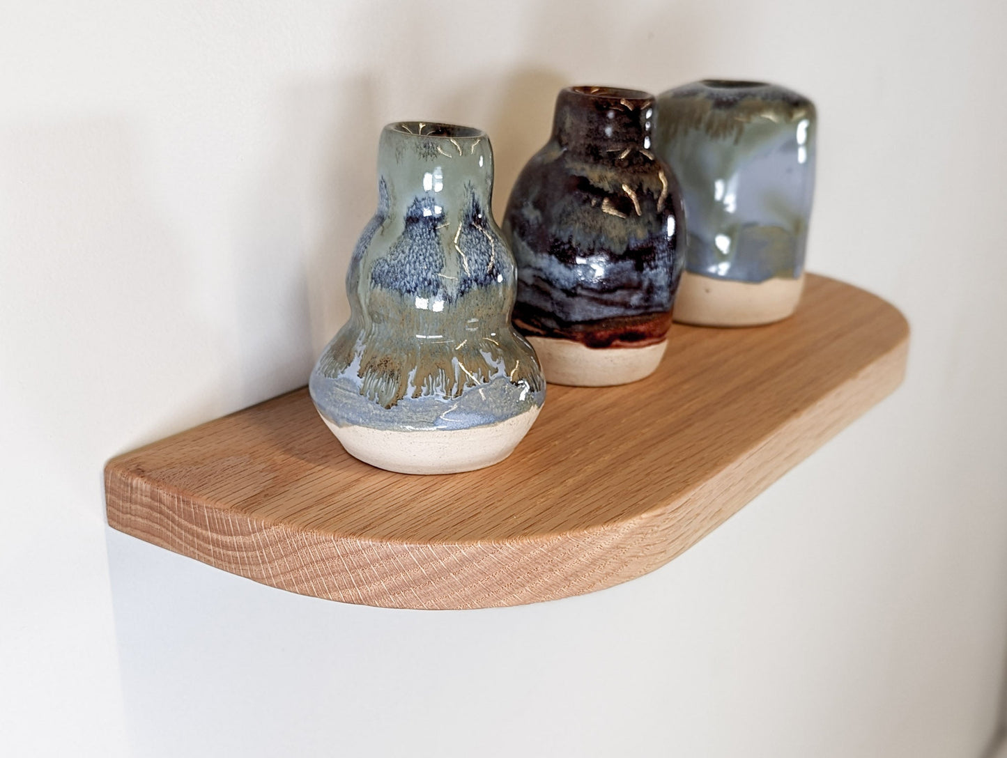 Angled view of a thin oak floating shelf with rounded corners and three vases resting on top