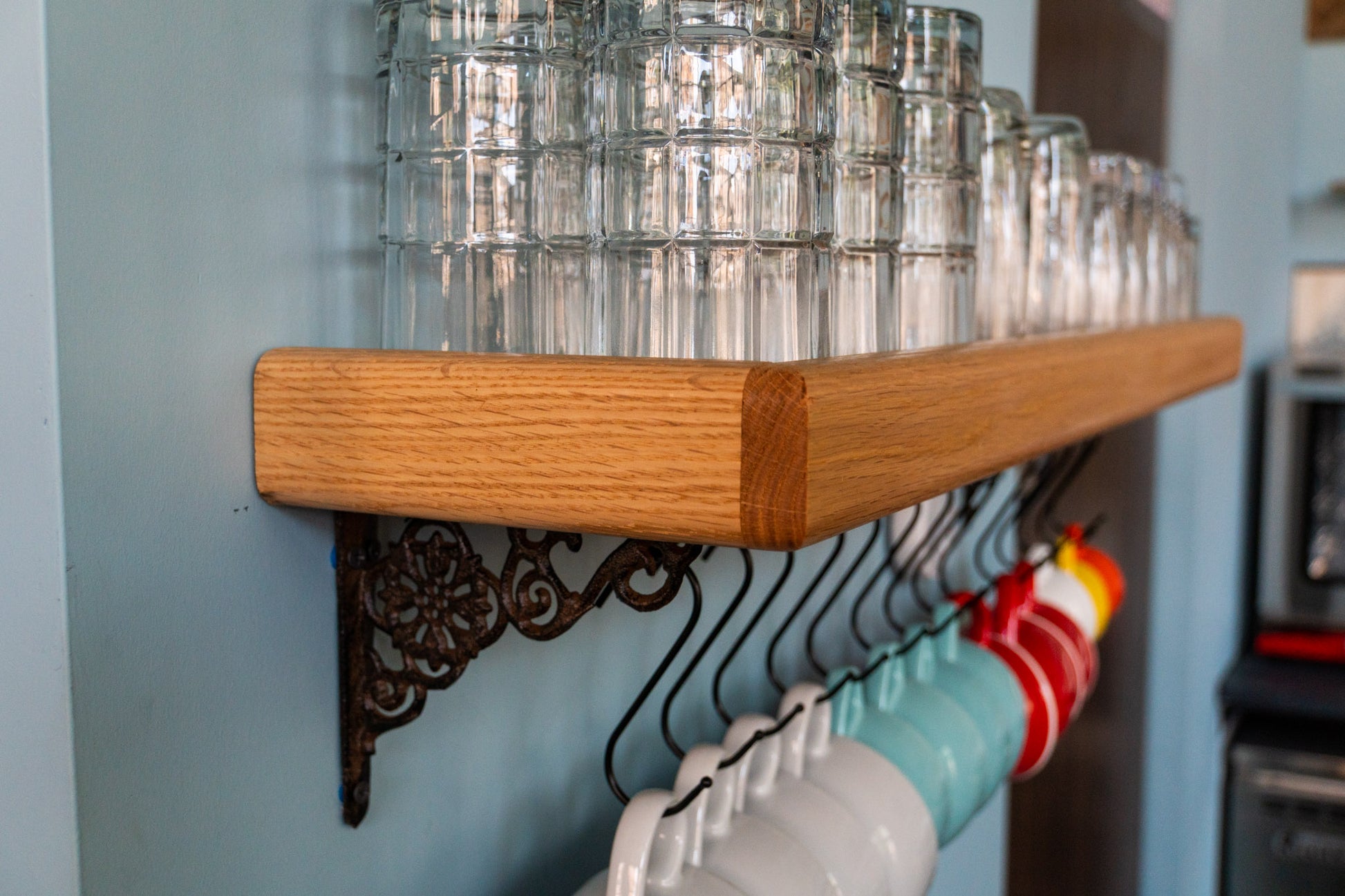 The side view of the Customizable Coffee Mug Shelf. The oak and mahogany wood shelf has an edge that rises upward creating a secure holding for stacked glasses. The cast iron bracket features a whirling design and a variety of black metal hooks hang below the shelf holding white, blue, red, yellow, and orange coffee cups. 