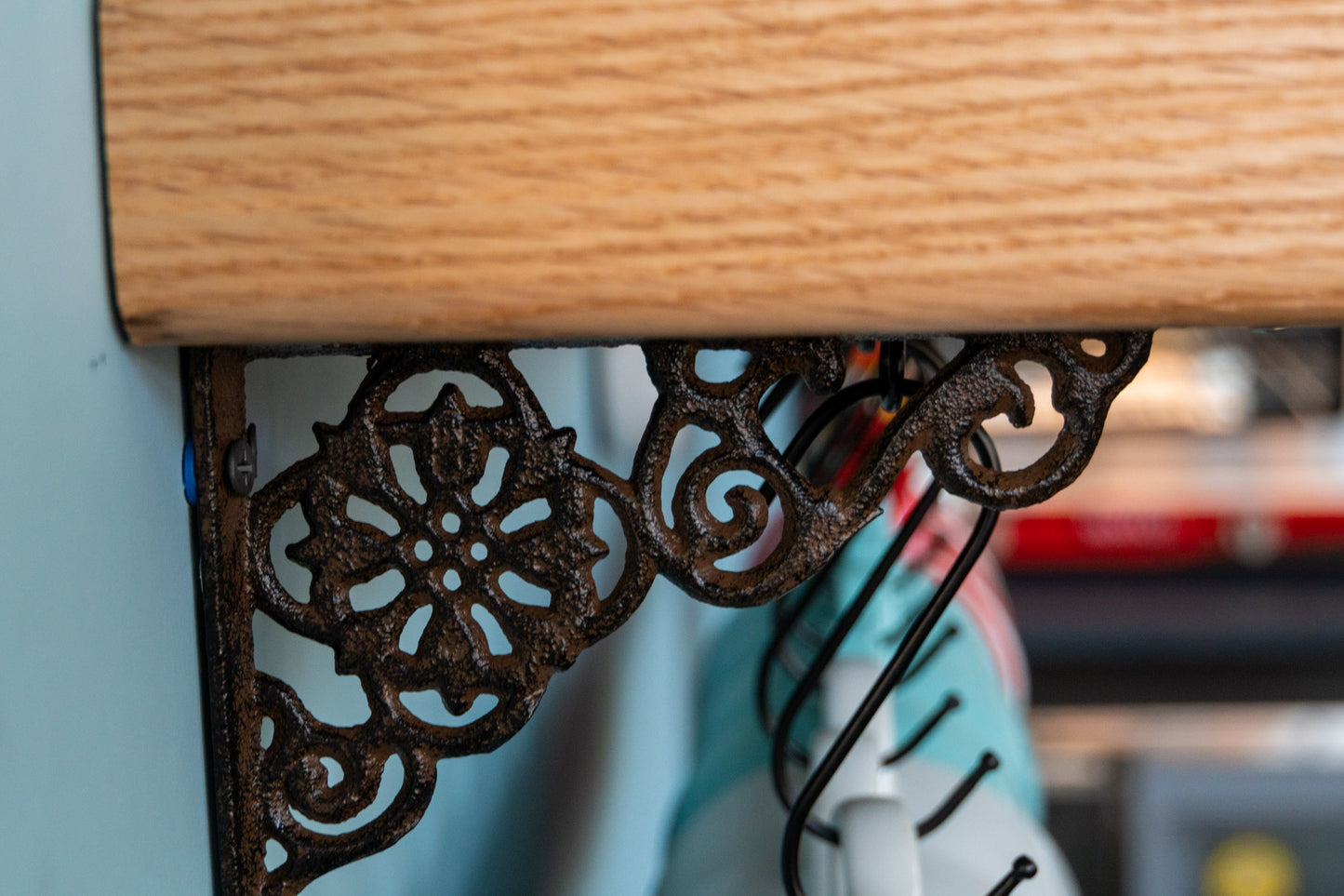 A close-up image of a cast iron bracket. The bracket is intricately designed with whirling motions. An eight-petaled flower set on a four-arched circle is in the center. The bracket holds up the oak shelf and black hooks dangle from below the shelf. Coffee cups are just out of focus. 