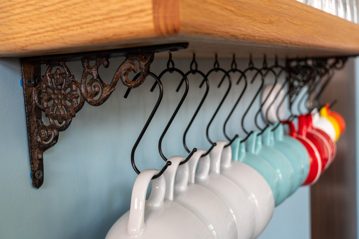 An intricately designed cast-iron bracket holds an oak shelf and is wall mounted to a blue wall. S-shaped metal hooks are attached to the bottom of the shelf and hold a variety of coffee mugs in white, blue, red, yellow, and orange colors.