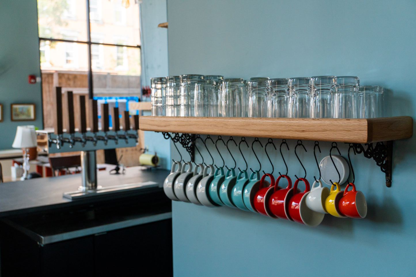 A high-quality and customizable coffee mug shelf is mounted on a blue wall. Various sized glasses are stacked on top of the shelf and below hang five white mugs, four blue mugs, three red mugs, and a yellow and orange espresso cups. In the back, out of focus is a bar tap with seven wooden taps in descending sizes.