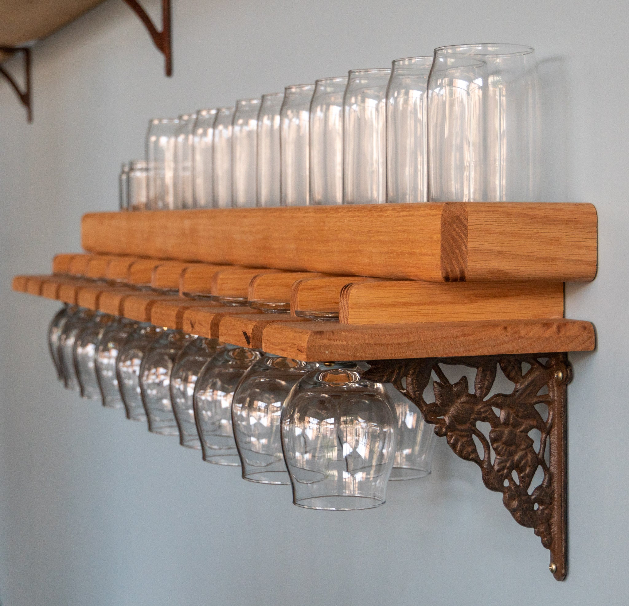 A side view of our Oak or Mahogany Beer Glass Shelf. The oak shelf holds 13 drink glasses securely as a variety of tulip glasses hang upside down below. A cast-iron bracket features a floral pattern. The shelf is mounted on a blue wall.