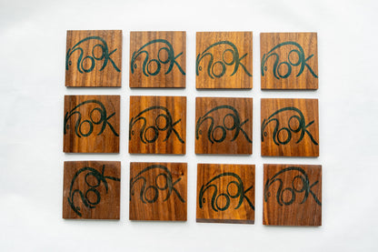 Three rows of four NookWoodworking coasters are viewed from above. These solid wood coasters are square with the word, "nook" stamped on them in large, rounded, blue, letters.