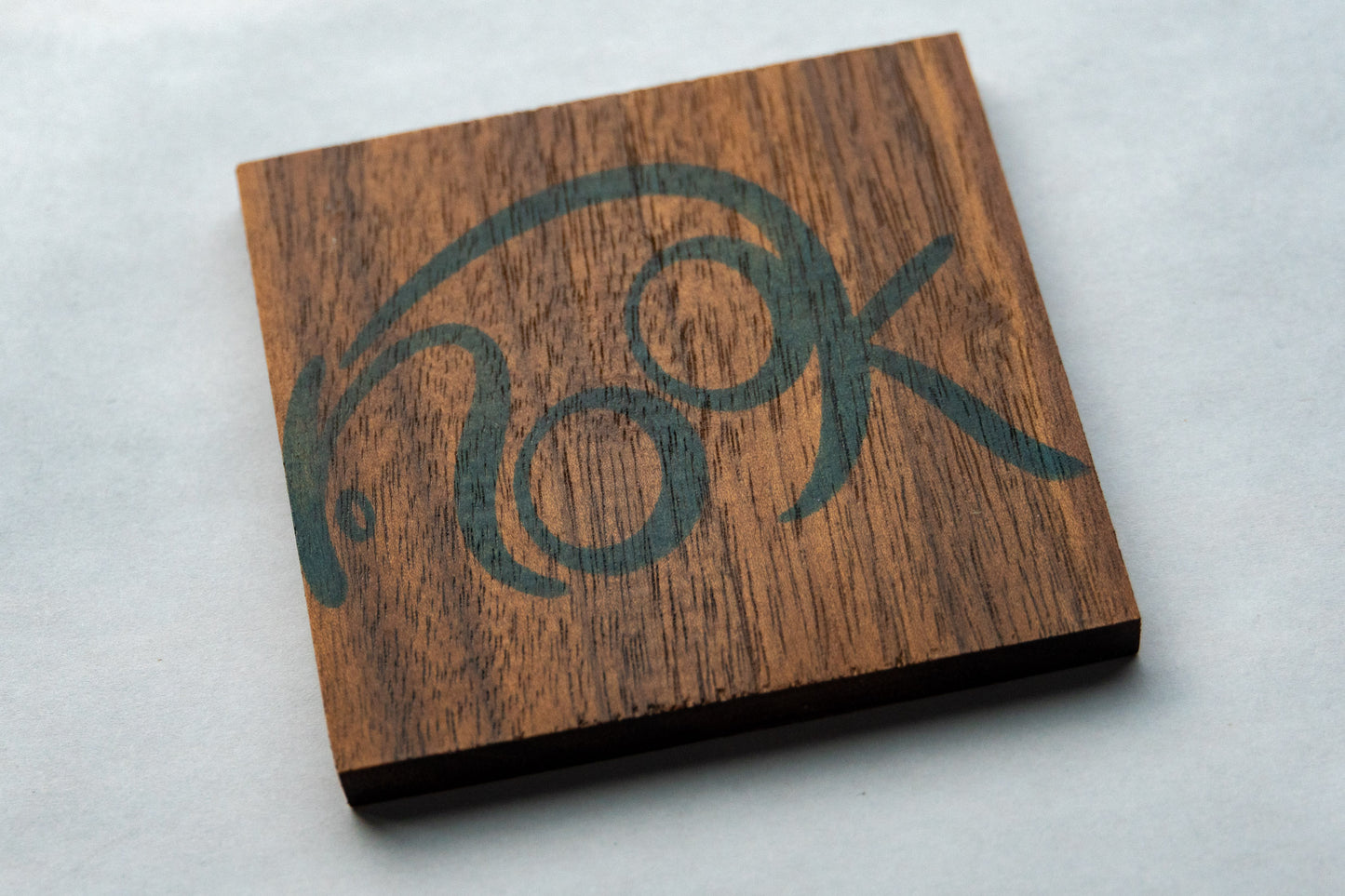 A square walnut wooden coaster, with the word nook stylistically written with large rounded letters in blue in.
