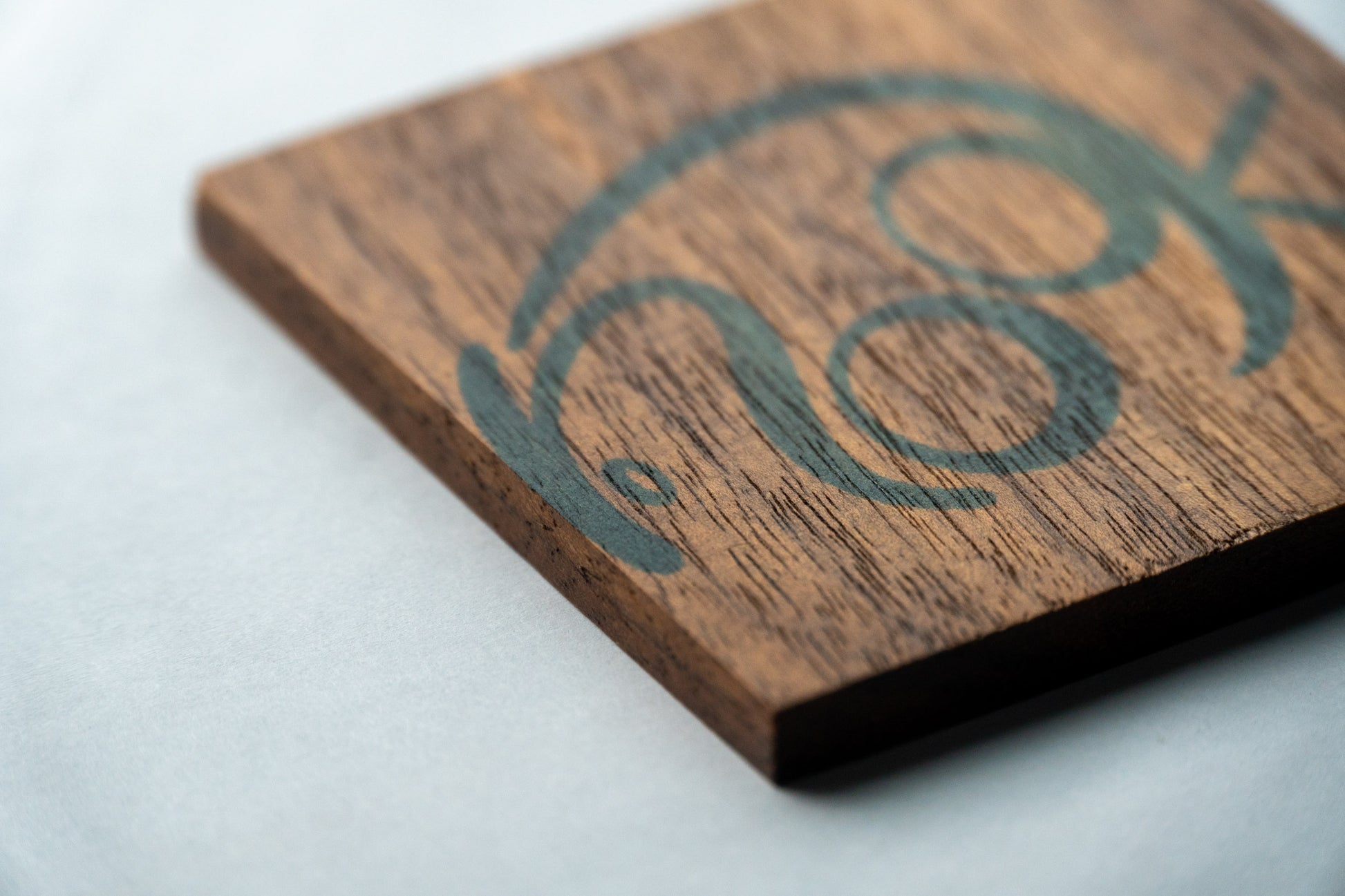 A close-up of the corner edge of Nook wooden coasters. The solid walnut coaster is square with sharp edges. The word "nook" is stamped on the top and just out of focus.