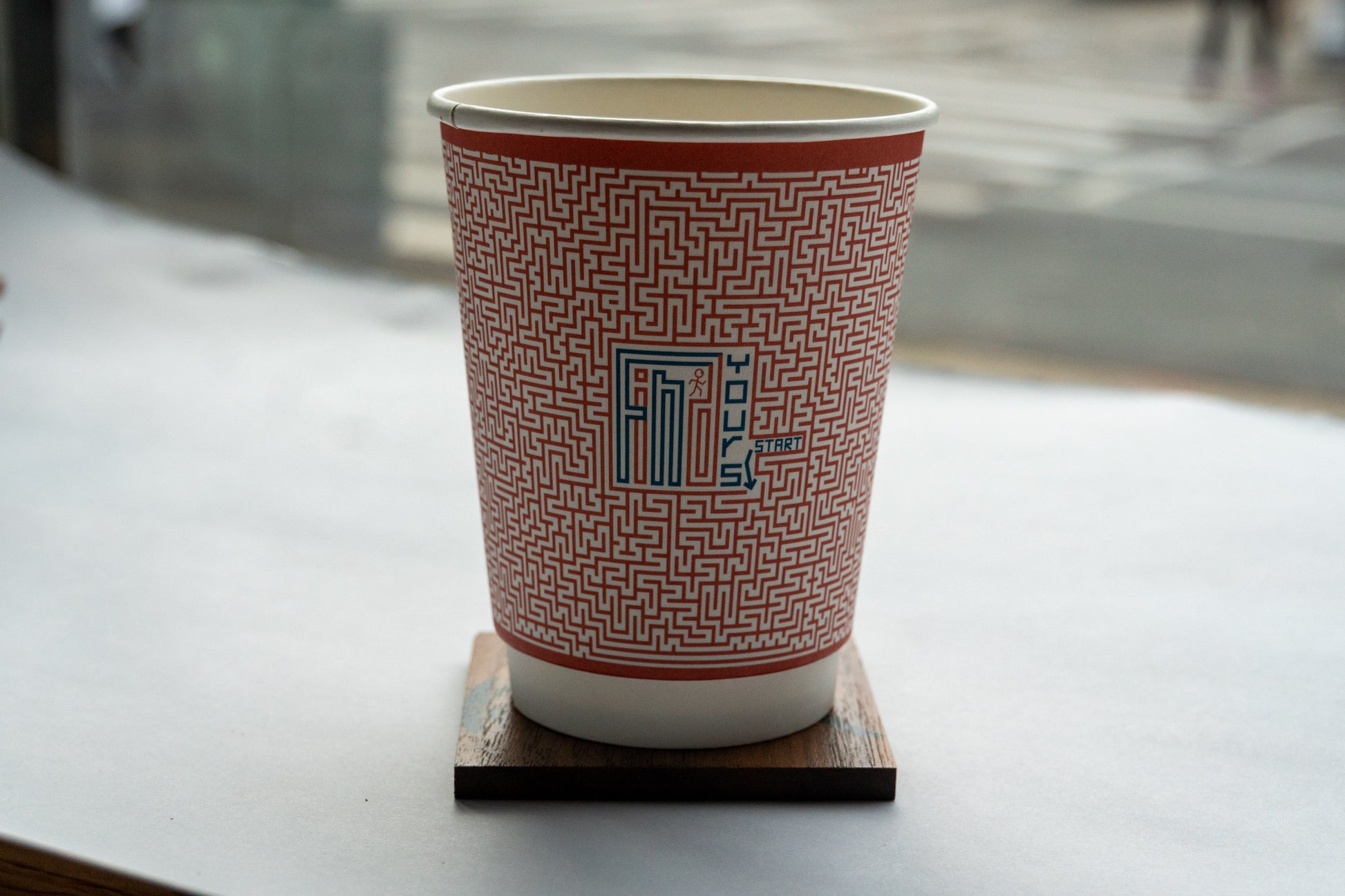 A coffee cup sits on a wooden coaster. The coffee cup features a red maze like design with the words "Find Yours" in alternating shades of red and blue. The word, "Start" is completely blue and an arrow points to the beginning of a maze. A stick figure is perched on top of the "D" in Find. 