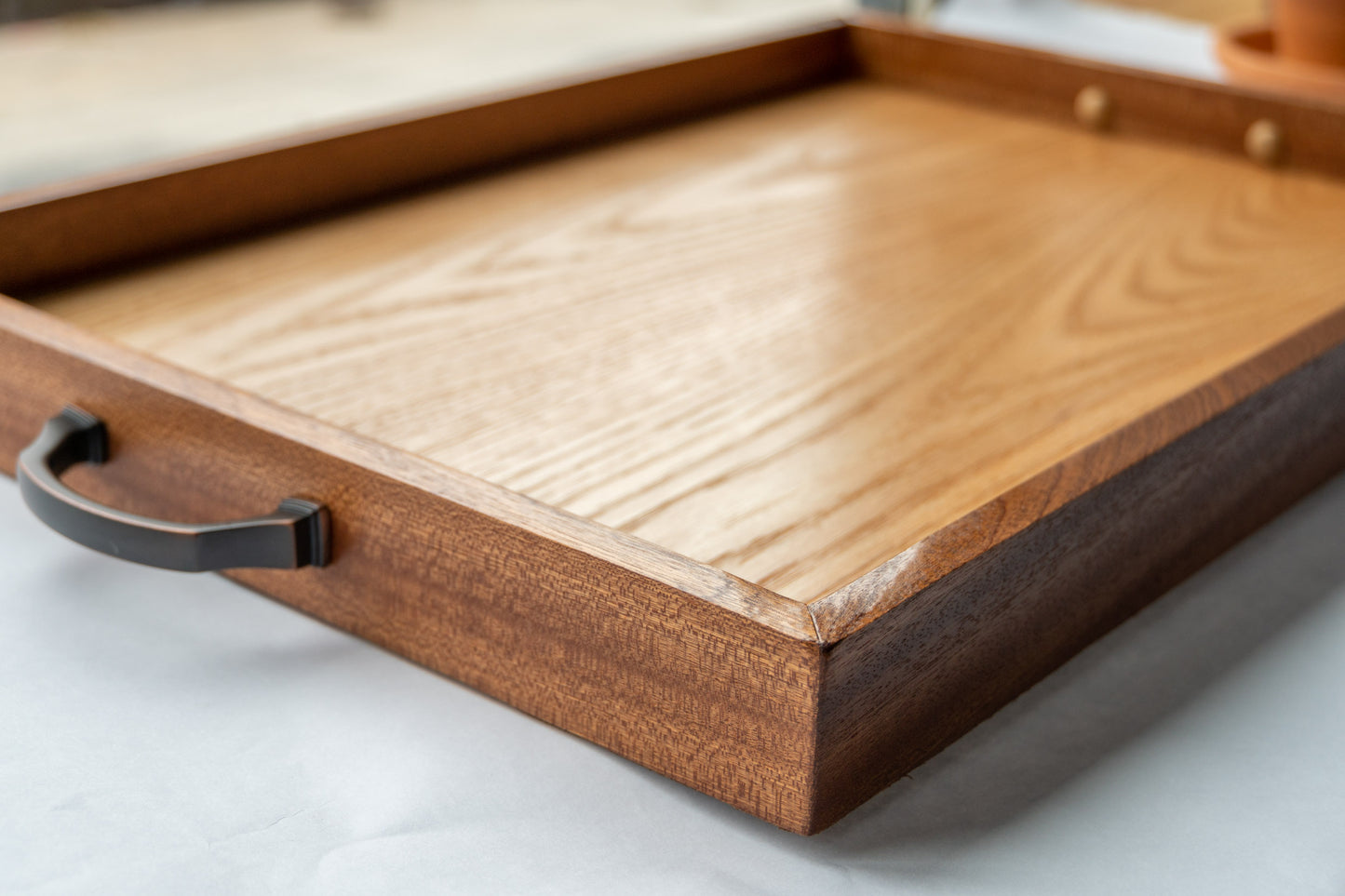 A side view of our Luxury Serving Tray. Mahogany wood forms the raised lip of the tray in an attractive dark brown grain. Inside, gleaming oak wood serves as the base. On either side of the tray a comfortable brass handle protrudes. The tray sits on a white surface.