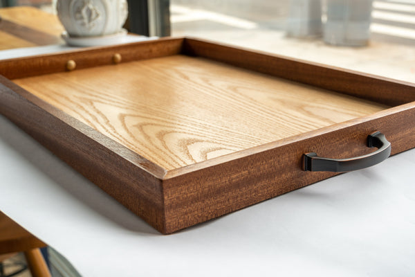 A side view of our Luxury Serving Tray. Mahogany wood forms the raised lip of the tray in an attractive dark brown grain. Inside, gleaming oak wood serves as the base. On either side of the tray a comfortable brass handle protrude. The tray sits on a white surface. Just in the back of the frame sits an out-of-focus ornate pot.